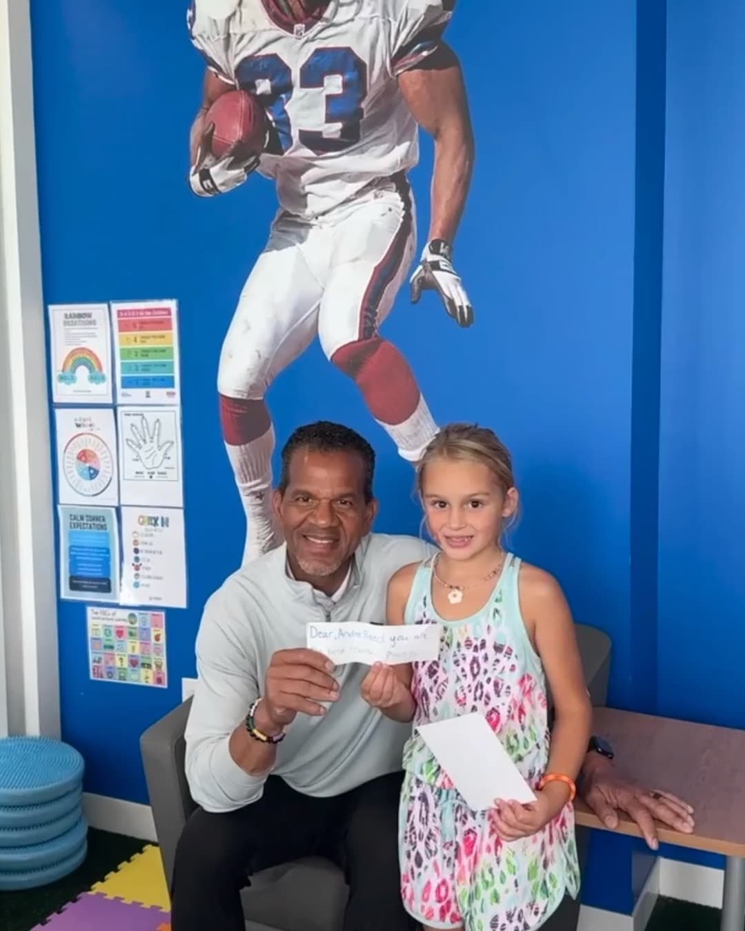 Andre Reed seated with a girl holding a handwritten thank-you note, in front of a Bills #83 cutout in a reading room