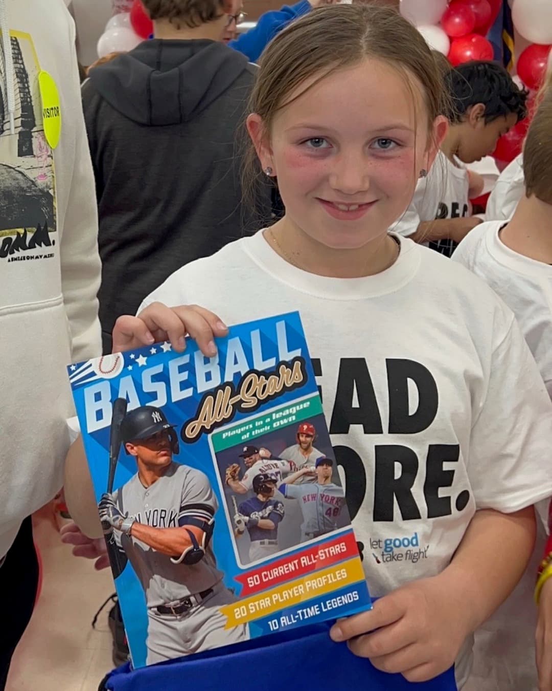 Girl holds a Baseball All-Stars book under a balloon arch at a book launch