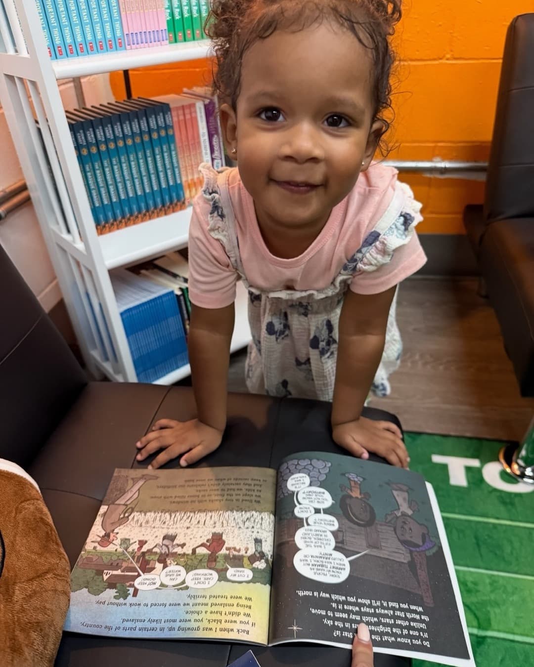 A toddler leaning over an open picture book in a reading room lined with full bookshelves