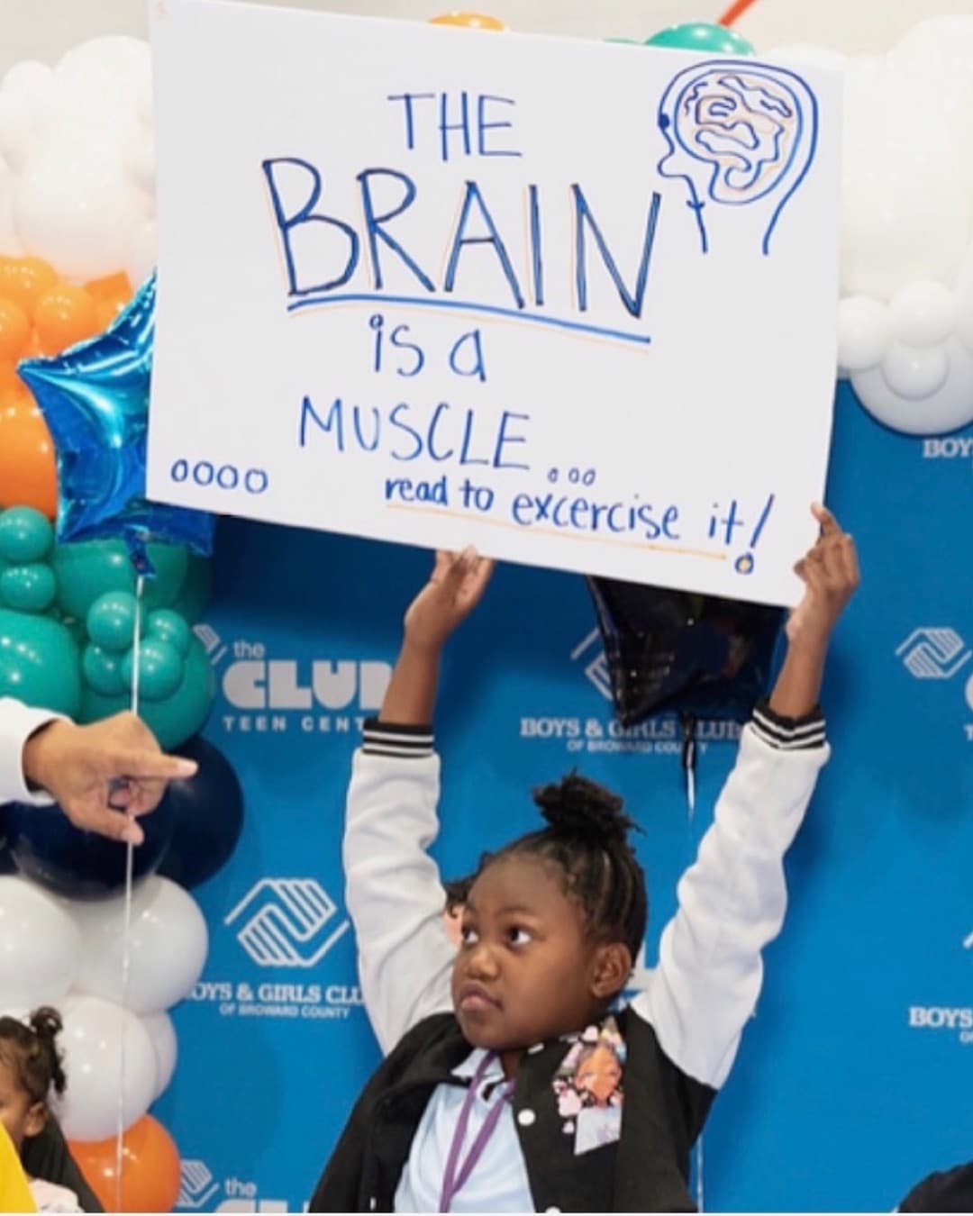 A girl holding a hand-lettered sign that reads "The brain is a muscle — read to exercise it!" at a Boys & Girls Club event