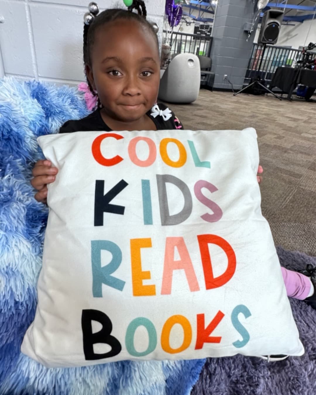 A girl in a Read with Reed reading room holding up a "Cool Kids Read Books" pillow, sitting on a fuzzy blue beanbag