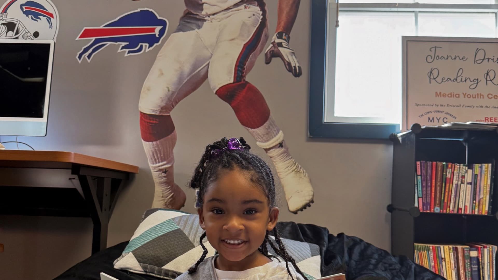 A girl reads a picture book beneath a large Andre Reed #83 mural in a Boys & Girls Club reading room