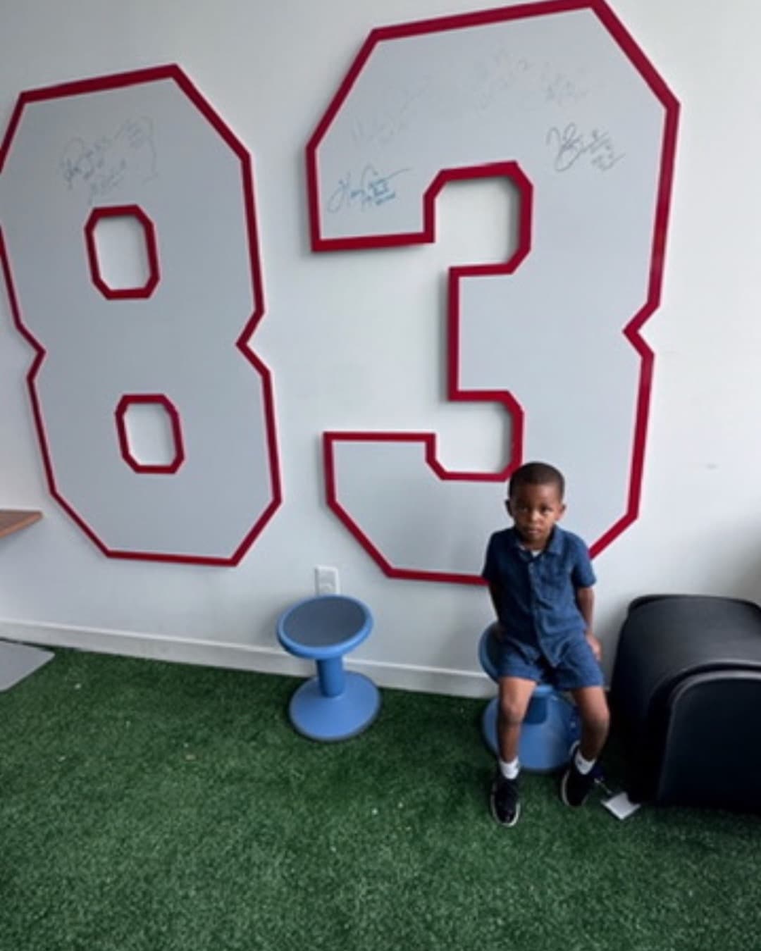 A boy seated on a wobble stool in front of a large signed "83" jersey-number wall decal on turf-green flooring