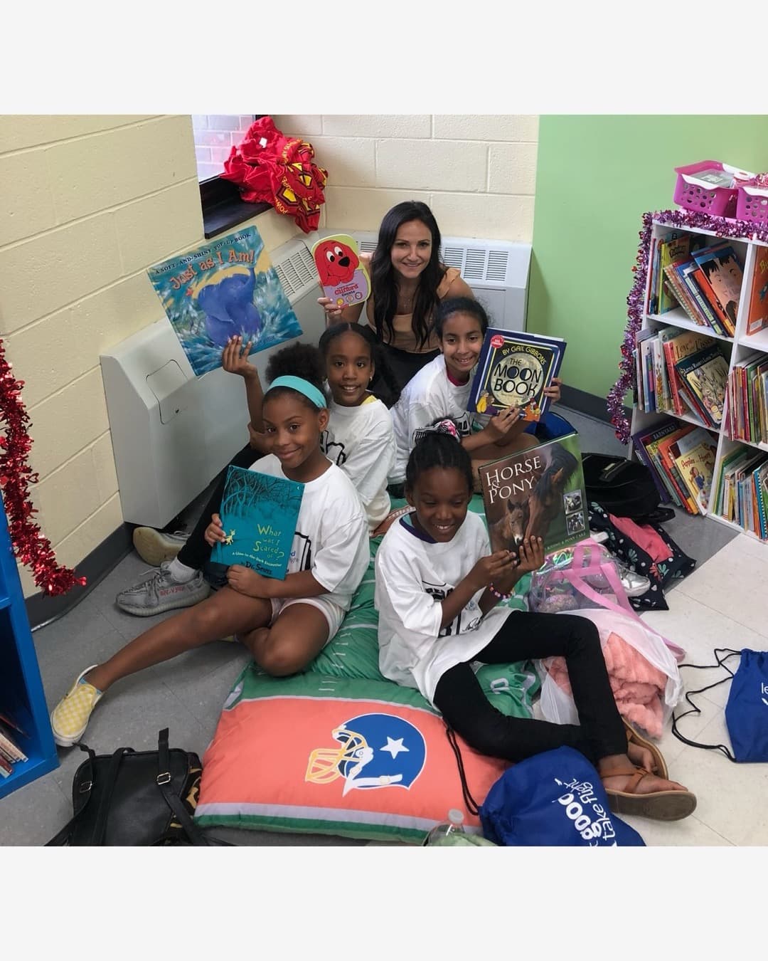 A group of girls reading books on floor cushions with a mentor in a reading room stocked with bookshelves