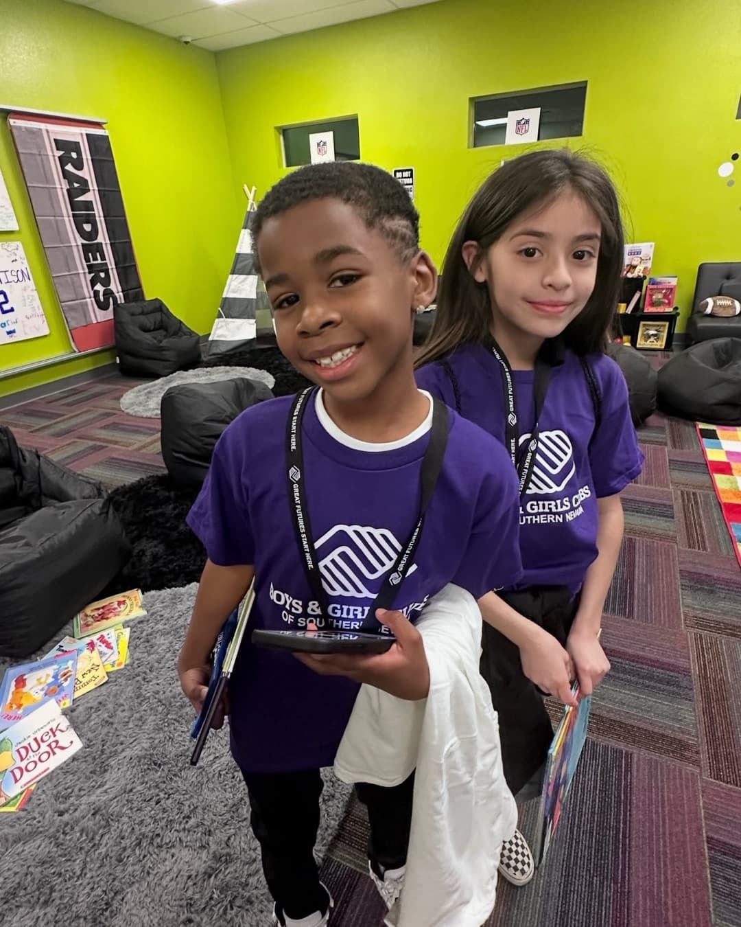 Two kids in Boys & Girls Club shirts smile with their books in a reading room