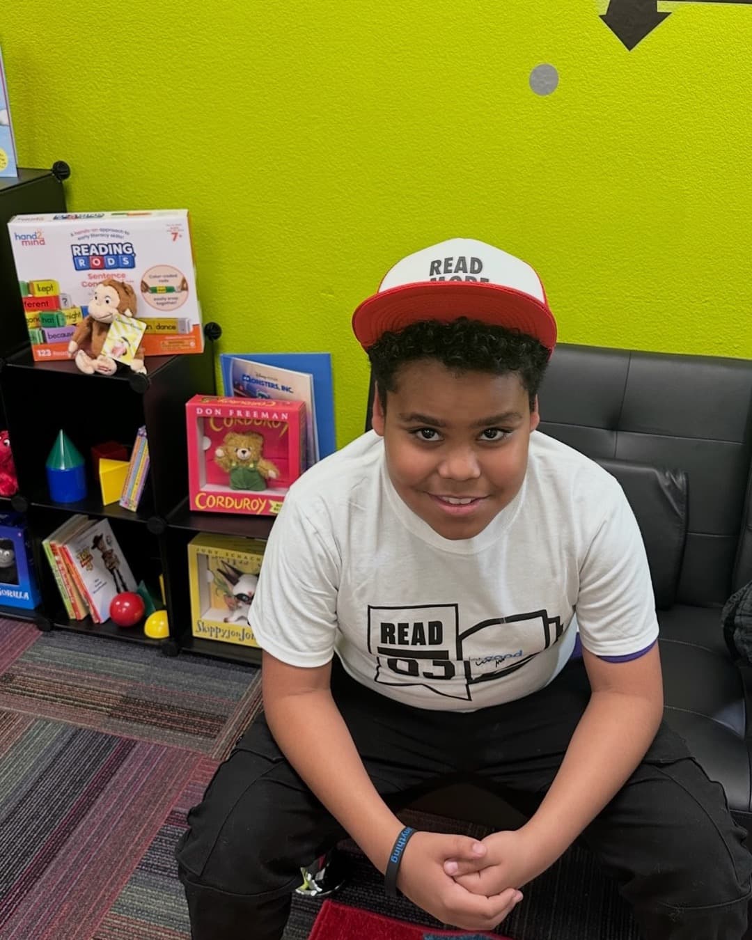 A boy wearing a "Read More 83" cap and a Read 83 t-shirt in a reading nook stocked with picture books and Reading Rods